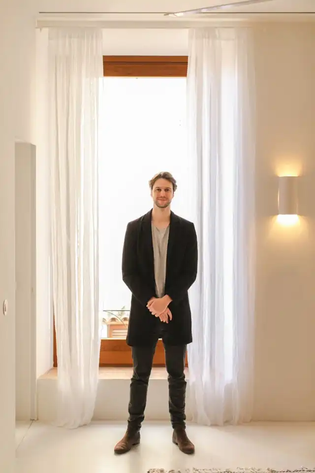 Andrew standing in front of a tall window framed by white curtains