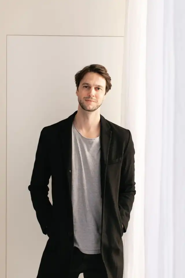 Andrew Nocker in a black coat and grey t-shirt, standing by a white linen curtain